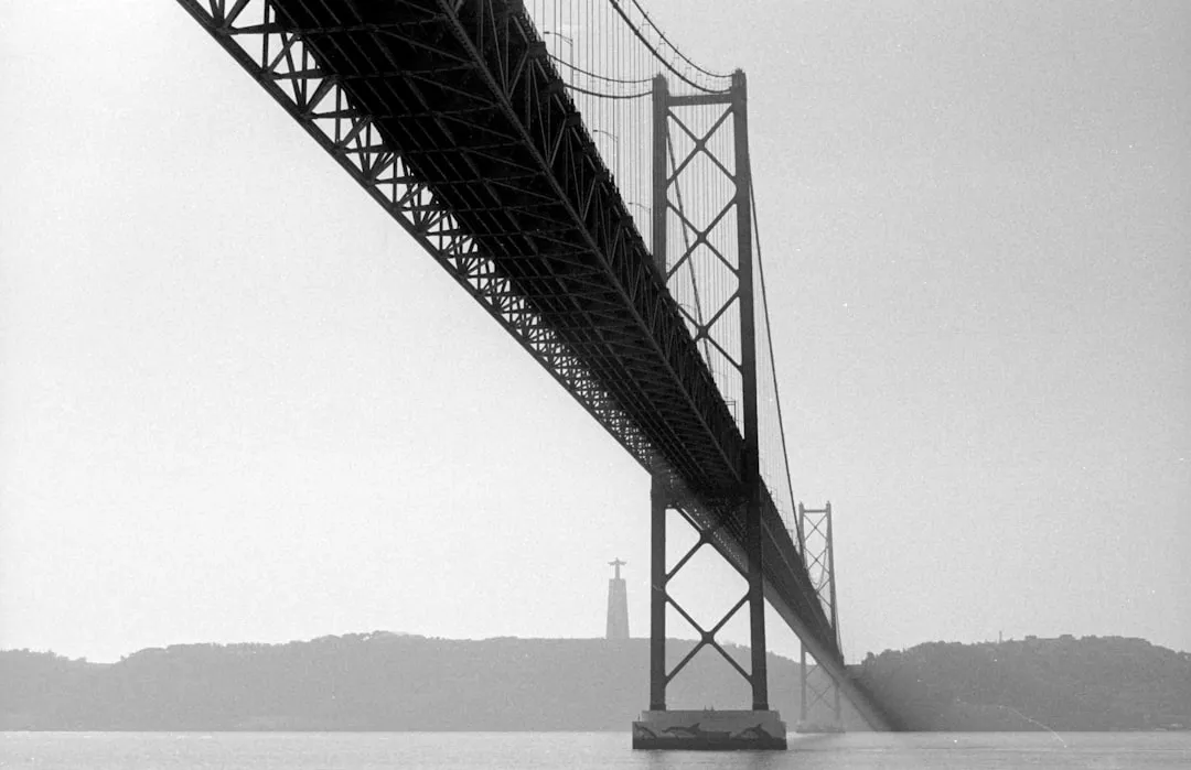 A black and white photograph of the 25 de Abril Bridge in Lisbon, Portugal, spanning over the Tagus River with a lighthouse in the background.